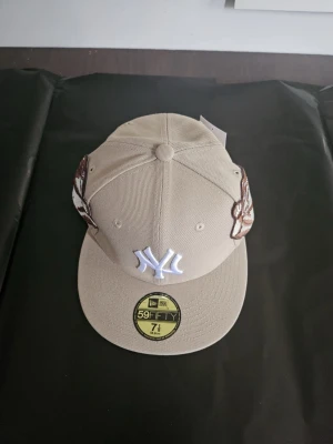Beige New Era 59FIFTY keps NY - Snygg beige keps från New Era med NY-logga framtill och broderad konst på sidorna. Modellen är 59FIFTY med platt skärm och klassisk passform. Perfekt för dig som vill sticka ut med coola detaljer och streetstyle-vibe.