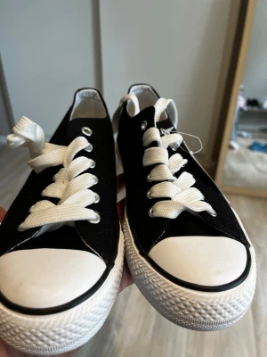 Svarta låga sneakers med vit sula - Svarta sneakers med klassisk låg profil, vita gummisulor och tåhätta samt vita skosnören. Skorna har en tidlös design med canvasmaterial och svarta detaljer längs sulan. Perfekta för en avslappnad och stilren look.