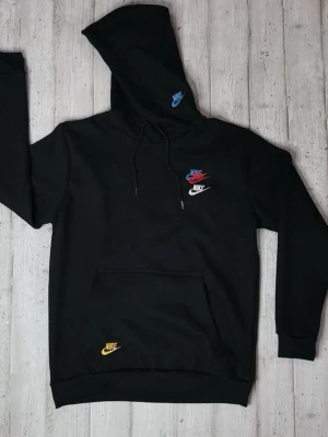 Svart Nike hoodie med färgglada loggor - Svart hoodie från Nike med flera broderade loggor i blått, rött, vitt och gult. Klassisk känguruficka framtill, justerbar huva och ribbade muddar. Perfekt för en avslappnad streetwear-look.