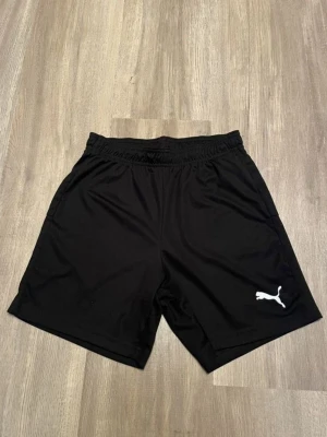 Svarta shorts från Puma - Svarta sportiga shorts från Puma med elastisk midja och vit logga på ena benet. Perfekta för träning eller chill, tillverkade i ett lätt och bekvämt polyestermaterial.