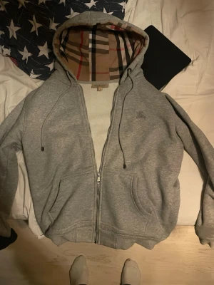 Grå zip-hoodie från Burberry - Snygg grå hoodie med dragkedja från Burberry. Klassisk rutmönstrad huva och broderad logga på bröstet. Mjuk bomullskänsla, två fickor fram och justerbara snören i huvan. Perfekt för en chill och stilren look.