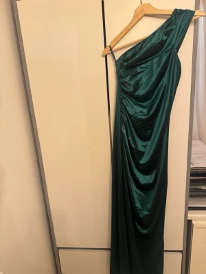 Grön långklänning i satin oneshoulder - Snygg mörkgrön långklänning i glansigt satinmaterial med oneshoulder-design och draperad effekt. Klänningen har en elegant slits på sidan och sitter figurnära upptill. Perfekt för bal eller festkvällar.