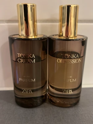 Zara Tonka Cream & Obsession Parfym - Två eleganta parfymflaskor från Zara: Tonka Cream och Tonka Obsession. Flaskorna är cylinderformade i brunt glas med guldfärgade lock och text. Perfekta för dig som gillar lyxiga och moderna dofter med stilren design.