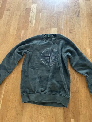 Camo crewneck från Stone Island - XL men är som L!                                               Snygg camouflagemönstrad crewneck från Stone Island med stort broderat kompassmärke på bröstet. Tröjan är i gröna toner, har rund hals och ribbade muddar. Perfekt för dig som gillar streetwear och vill sticka ut med en ikonisk logga.