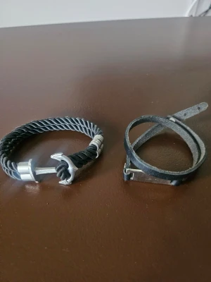 Svarta armband med ankardetalj - Två coola armband, ett med svart tvinnat rep och silverfärgad ankardetalj, och ett smalt svart läderarmband med enkel metallspänne. Perfekt för dig som gillar stilrena accessoarer med marint tema. Det med ankaret är endast använt ett par ggr