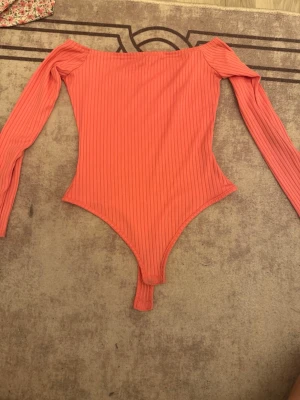 Ribbad korallfärgad body NLY One - Trendig ribbad body i korall från NLY One. Off shoulder-modell med långa ärmar och figurnära passform. Mjukt stretchigt material som sitter snyggt på kroppen. Perfekt att styla med jeans eller kjol för en cool look.