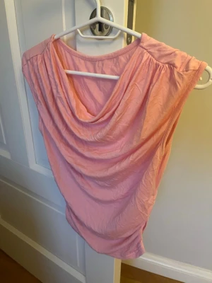 Ljusrosa draperad topp - Snygg ljusrosa topp med draperad framsida och breda axlar. Toppen är ärmlös och har en avslappnad passform, perfekt att styla med jeans eller kjol. Tillverkad i mjuk bomull som känns skön mot huden.