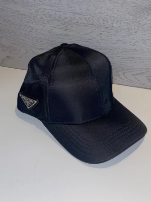 Svart keps från Prada i satin - Stilren svart keps från Prada i glansigt satintyg med klassisk böjd skärm och ikonisk metallogo på sidan. Kepsen har sex paneler och justerbar passform baktill. Perfekt accessoar för en trendig och lyxig look.