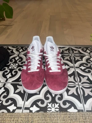 Vinröda Adidas Gazelle sneakers - Säljer ett par vinröda Adidas Gazelle sneakers i mocka med klassiska vita ränder på sidorna och vita snören. Skorna har låg profil och vit sula, samt Adidas-logga på plösen och hälen. Perfekta för dig som gillar retrostil och vill sticka ut.priset kan diskuteras 