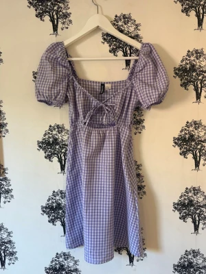 Lila rutig klänning med puffärmar - Superfin lila och vit rutig klänning från H&M med puffärmar och söt knytning framtill. Klänningen har en kort längd och är gjord i bomull, perfekt för vår och sommar. Den har en feminin v-ringning och sitter normalt på kroppen.