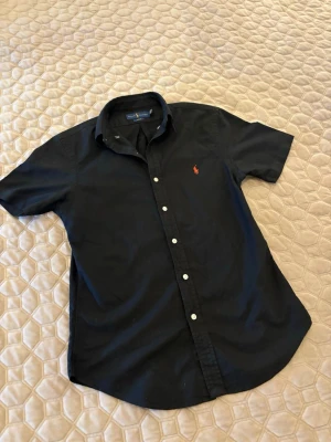 Svart kortärmad skjorta från Ralph Lauren - Svart skjorta från Ralph Lauren med korta ärmar och klassisk krage. Skjortan har vita knappar och en röd broderad logga på bröstet. Tillverkad i bomull och har en stilren, tidlös look.