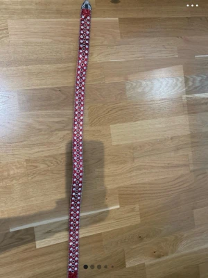 Rött glittrigt bälte med nitar - Snyggt rött bälte med glittrig yta och silverfärgade nitar längs hela bandet. Spännet är dekorerat med små stenar för extra bling. Perfekt accessoar för att lyfta din outfit med lite extra edge.
