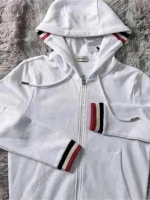 Vit hoodie från Moncler med dragkedja - Vit hoodie från Moncler med hel dragkedja och huva. Ärmsluten och insidan av huvan har snygga ränder i rött, svart och beige. Tillverkad i mjukt material, perfekt för en chill och stilren look.