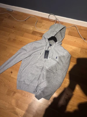 Grått loungewear-set från Polo Ralph Lauren - Säljer ett grått loungewear-set från Polo Ralph Lauren med hoodie och matchande mjukisbyxor. Setet har dragkedja på tröjan, fickor och elastisk midja på byxorna. Klassisk Polo-logga broderad på byxan. Perfekt för chill dagar hemma. Eller om du vill ta det lite mer chill ute, wjir snygg dress helt ny aldrig använd! Skriv vid funderingar osv. det är storlek ”S” tröjan!!!!