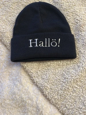 Mörkblå mössa - Snygg svart mössa i ribbstickad bomull med uppvikt kant och vit broderad text 'Hallö!' framtill. Perfekt accessoar för att hålla värmen och addera lite attityd till din outfit.