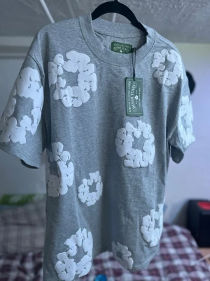 Grå t-shirt från Denim Tears - Snygg grå t-shirt från Denim Tears med vita blommiga mönster över hela tröjan. T-shirten har rund halsringning och korta ärmar. Perfekt för dig som vill sticka ut med en unik och trendig look.