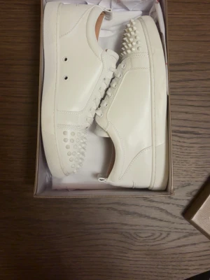 Vita sneakers från Christian Louboutin - Säljer ett par vita sneakers från Christian Louboutin med klassiska röda sulor och coola nitar på tån. Perfekta för dig som vill sticka ut med exklusiva detaljer. Storlek 42 och bara använda ett fåtal gånger. Hör gärna av dig vid funderingar.  