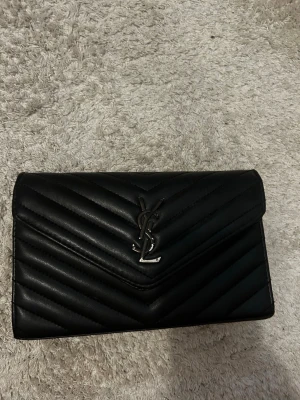 Svart YSL väska - Snygg svart väska. Kommer med kedja. Loggans färg har skrapats bort på några ställen men annars toppenskick och har även ett fack i väskan ❤️