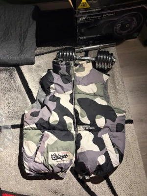 Camo dunväst med Chicago-tryck - Snygg dunväst i camomönster med stora broderade Chicago-detaljer på ryggen och mindre tryck framtill. Västen har dragkedja, två fickor och hög krage. Perfekt för dig som vill sticka ut med streetwear-stil.