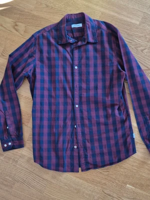 Rutig vinröd skjorta från Jack & Jones - Snygg långärmad skjorta från Jack & Jones vinrött och mörkblått rutigt mönster. Skjortan har klassisk krage, knäppning framtill och är tillverkad i mjuk bomull. Perfekt för en avslappnad och trendig look.