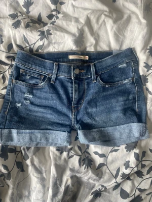 Levi's blå jeansshorts med upprullad kant - Säljer ett par klassiska blå jeansshorts från Levi's med upprullad kant och slitna detaljer framtill. Shortsen har fem fickor, bälteshällor och Levi's ikoniska läderpatch bak. Perfekta för varma dagar och enkel att matcha med allt.