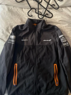 Svart McLaren jacka med orange detaljer - Säljer en svart McLaren jacka med dragkedja och orange detaljer vid fickorna. Jackan har tryckta loggor från olika sponsorer på bröstet och ärmarna. 