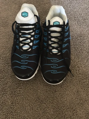 Nike Air Max Plus svarta/blå sneakers - Nike Air Max Plus sneakers i svart med blå detaljer och vit sula. Skorna har en sportig design med vågformade linjer och meshmaterial på ovandelen. Klassisk Air Max-dämpning i sulan och vita meshpartier på plösen och insidan. Perfekta för en streetwear-look.