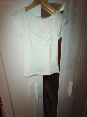 Vit topp med spets och rosett - Söt vit topp från Lager 157 med korta ärmar, v-ringning och spetsdetaljer längs urringningen. Liten rosett framtill och lätt rynk vid bysten ger en feminin touch. Perfekt att matcha med jeans eller kjol.