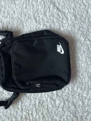 Svart Nike ryggsäck med justerbara remmar - En stilren svart Nike ryggsäck i slitstarkt tygmaterial med justerbara axelremmar och dragkedja framtill. Perfekt för dig som vill ha en enkel och praktisk väska till skolan eller stan. Ryggsäcken har en klassisk form och flera fack för enkel organisering.