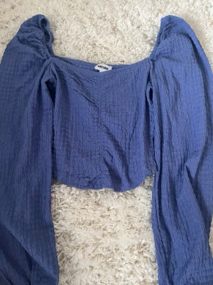 Blå croppad topp från Bubbleroom - Superfin blå croppad topp från Bubbleroom med långa puffärmar och fyrkantig ringning. Toppen är gjord i ett lätt, krinklat material som ger en cool struktur. Perfekt att styla med höga jeans eller kjol för en trendig look.