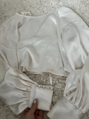 Vit satinsblus  - Snygg vit blus i satin med långa puffärmar och dekorativa knappar vid ärmsluten. Blusen har en öppen rygg med knytband upptill och en kort, rak passform. Perfekt för dig som gillar trendiga detaljer och en lyxig känsla.