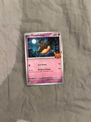 Pumpkaboo Pokémonkort - Pumpkaboo Pokémonkort med lila och svart design, motiv av Pumpkaboo under en fullmåne. Kortet har en orange Pikachu-stämpel och visar attackerna Seed Bomb och Reckless Charge. Perfekt för samlare eller Pokémon-fans.
