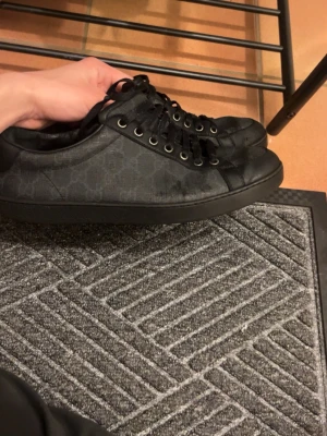 Svarta Gucci sneakers med monogram - Säljer ett par svarta Gucci sneakers med diskret GG-monogram på sidan. Skorna har svarta skosnören, metallöljetter och en låg profil. Materialet är en mix av läder och canvas, vilket ger en lyxig känsla och snygg look.