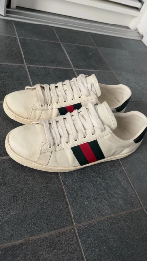  vita sneakers med ränder - Säljer ett par klassiska vita sneakers med gröna och röda sidoremsor. Skorna har lågt skaft, vita snören och är tillverkade i läder. Perfekta för dig som vill ha en stilren och ikonisk look.