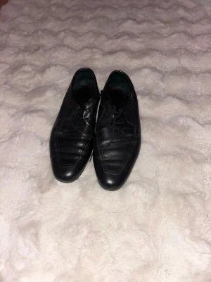 Svarta loafers i läder - Säljer ett par svarta loafers med klassisk design och snörning. Skorna är tillverkade i läder med diskreta sömmar och har en låg sula. Perfekta för dig som vill ha en stilren och tidlös look.