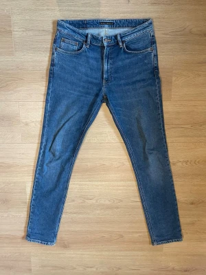 Blå slim fit jeans från Nudie - Snygga blå slim fit jeans från Nudie med klassisk femficksdesign och gul söm på bakfickorna. Jeansen har normal midja, dragkedjegylf och är tillverkade i bomull med en lätt stretch för extra komfort.                                                                                   Köpta i nudie jeans affär i Stockholm. Ej använda och ej tvättade.                                                                   Hör av dig vid frågor, pris kan diskuteras vid snabb affär 