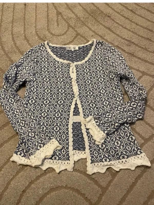 Blåvit mönstrad kofta med volangkant - Supersöt blåvit kofta med stickat mönster och volangkant. Koftan har rund halsringning, långa ärmar och vita virkade detaljer längs kanterna. Perfekt för dig som gillar en unik och bohemisk stil.