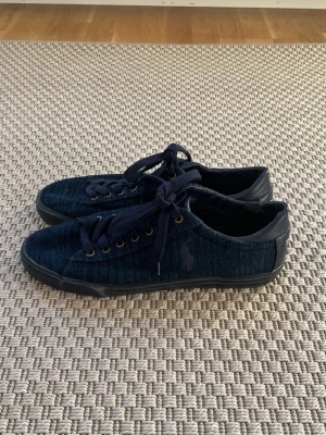 Mörkblå sneakers från Polo Ralph Lauren - Snygga mörkblå sneakers från Polo Ralph Lauren med denimliknande tyg och mörkblå läderdetaljer vid häl och snörning. Klassisk låg modell med svarta gummisulor och mörkblå skosnören. Perfekta för en stilren och avslappnad look. Storlek EUR 44