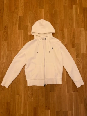 Vit hoodie från Polo Ralph Lauren - Snygg vit hoodie från Polo Ralph Lauren med dragkedja framtill och klassisk huva med snörning. Ikoniska lilla loggan broderad på bröstet. Två fickor framtill och ribbade muddar vid ärmar och midja. Perfekt för en clean och stilren look. QR kod finns i hoodien vilket bevisar att den är äkta. Pris kan diskuteras 