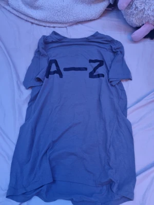 Grå t-shirt från A-Z med tryck - Säljer en grå t-shirt från A-Z med svart tryck på bröstet. T-shirten har klassisk passform och är tillverkad i mjuk bomull som känns skön mot huden. Perfekt till jeans eller shorts för en enkel och clean look.
