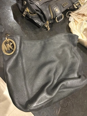 Svart Michael Kors handväska i skinn - Snygg svart handväska från Michael Kors i mjukt skinn med guldfärgade detaljer och stort MK-hänge. Väskan har flera innerfack med dragkedja och rymligt huvudfack. Perfekt för dig som gillar klassisk och stilren design.