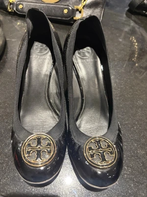 Svarta klackar från Tory Burch - Snygga svarta klackar från Tory Burch i lackat material med kilklack. Skorna har rund tå och är dekorerade med ett stort guldfärgat metallogo framtill. Klassisk och trendig design som passar till många olika stilar.