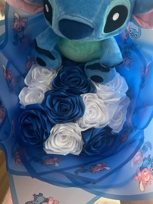 Blå Stitch-bukett med rosor - Söt bukett med en blå Stitch-gosedjur och konstgjorda rosor i blått och vitt. Buketten är inslagen i blått papper med Stitch-motiv och rosa detaljer. Perfekt som present till någon som gillar Disney eller söta dekorationer.