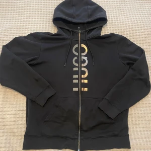 Svart hoodie med guldtryck från Hugo - Snygg svart hoodie från Hugo med dragkedja och stor huva. Framtill finns ett vertikalt guldigt logotryck som ger en exklusiv känsla. Tillverkad i mjukt bomullsmaterial och har två fickor framtill samt justerbara snören i huvan.