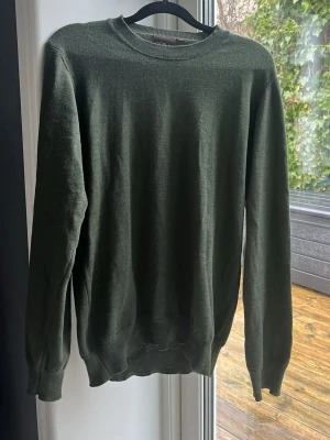Olivgrön stickad tröja Oscar Jacobson - Snygg olivgrön stickad tröja från Oscar Jacobson i tunn ullmix. Tröjan har rund halsringning, långa ärmar och ribbade muddar vid ärmslut och nederkant. Perfekt för lager på lager och stilrena outfits. Hör av dig vid minsta fundering. 