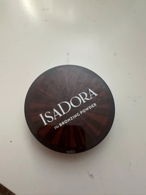 Isadora Bronzing Powder bronzer - Snygg bronzer från Isadora i en rund, brun plastförpackning med spegel i locket. Pudret har en varm, gyllenbrun nyans och är pressat med ett diskret mönster. Perfekt för att ge ansiktet en solkysst look.