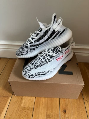 Yeezy Boost 350 V2 Zebra sneakers - Snygga Adidas Yeezy Boost 350 V2 'Zebra' sneakers med svartvitt zebramönster, röda detaljer och vit sula. Ovandelen är i stickad mesh för en skön och flexibel känsla. Ikonisk design som sticker ut och passar perfekt till streetwear.