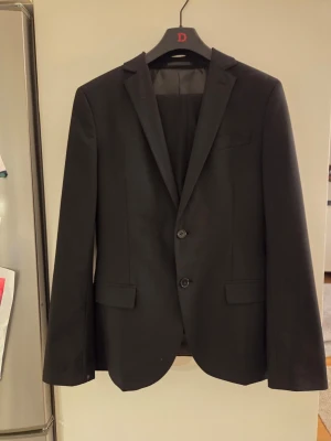 Svart kostym från Dressmann Slim Fit inkl vit skjorta H&M - Stilren svart kostym från Dressmann i modellen Slim Fit. Kavajen har två knappar framtill, två fickor med lock och en bröstficka. Fodret är randigt och ger en snygg kontrast. Perfekt för dig som gillar en klassisk och modern look. Vitt bomulls skjorta från H&M