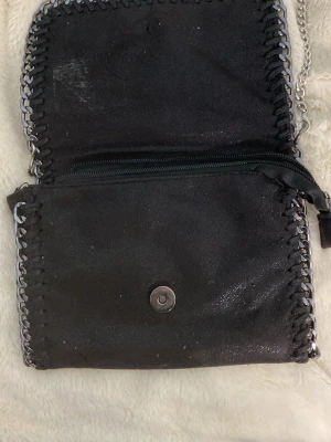 Svart axelremsväska med kedjedetaljer - Snygg svart handväska i mockaimitation med silverfärgade kedjedetaljer längs kanterna. Väskan har magnetlås, innerfack med dragkedja och axelrem i kedja. Perfekt för dig som gillar en edgy och trendig look.