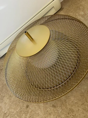 Guldig lampskärm i metallnät - Snygg lampskärm i guldigt metallnät med rund form och industriell känsla. Perfekt för att ge rummet en modern touch. Passar till taklampa och har ett fäste upptill för montering.